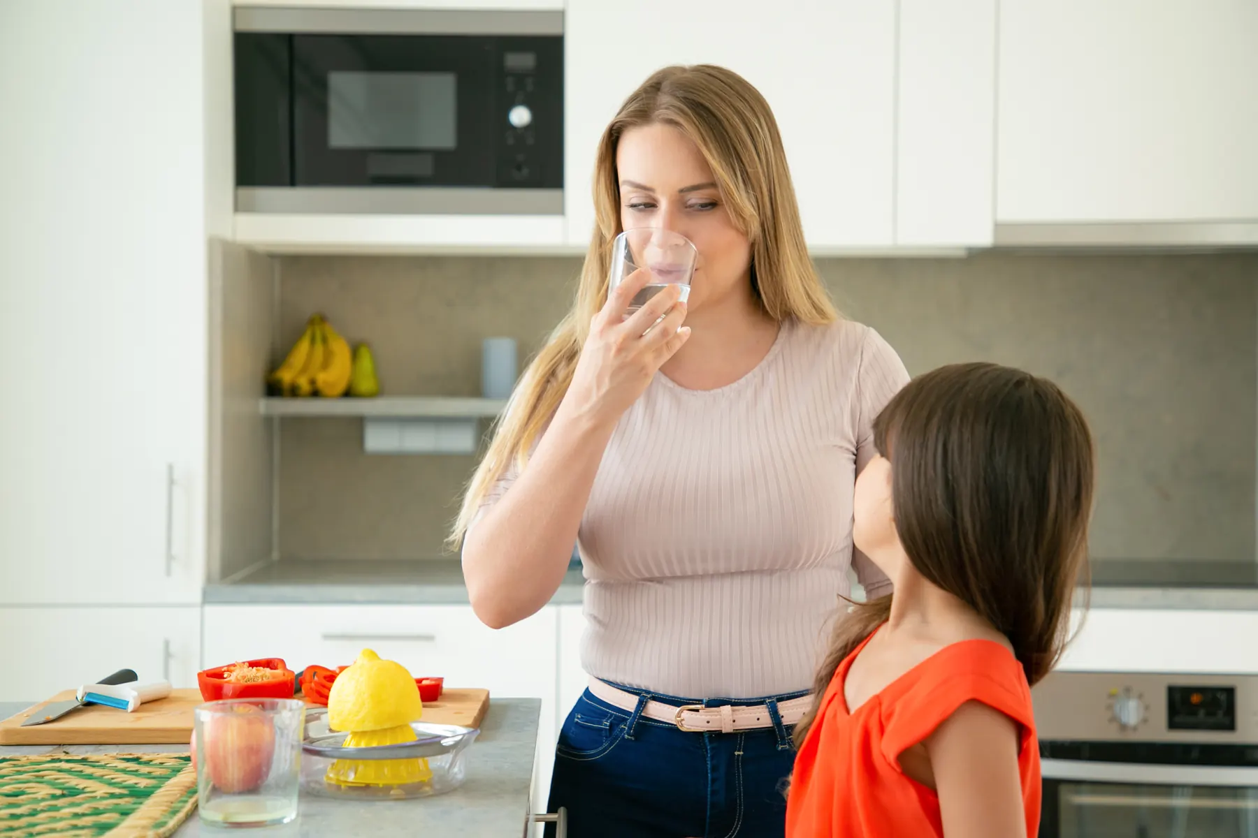 A woman sipping from a tall glass in a sunny kitchen while a girl looks on